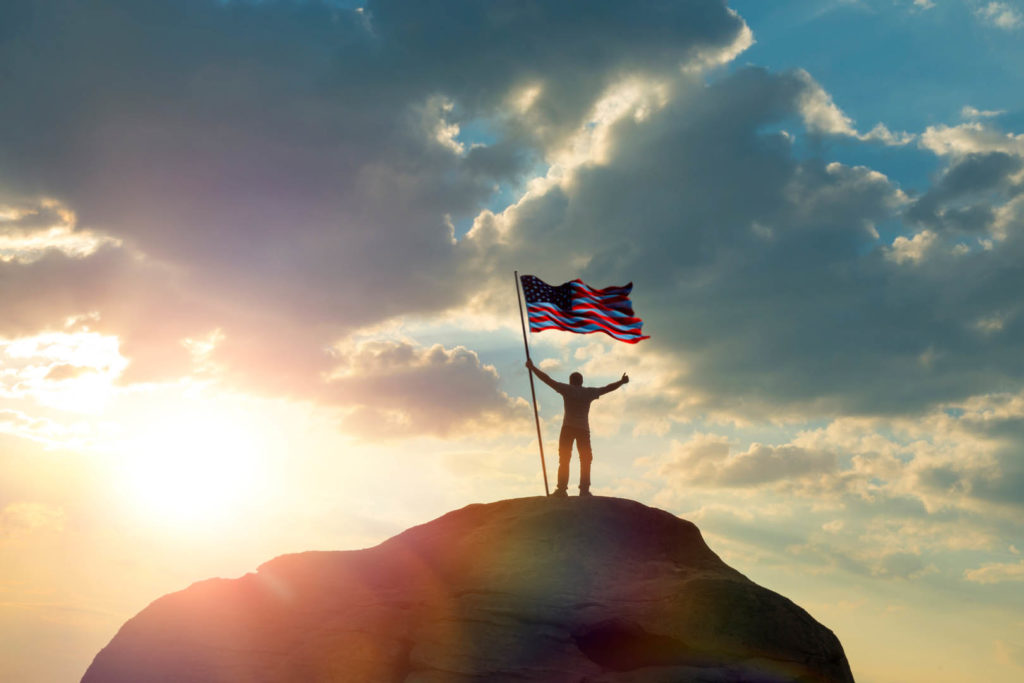 the man is standing on the top of the mountain, holding his hands up with the flag of the US America against the sunset. Business concept idea, success and achievements, career ladder, victory.