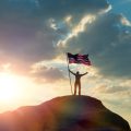 the man is standing on the top of the mountain, holding his hands up with the flag of the US America against the sunset. Business concept idea, success and achievements, career ladder, victory.